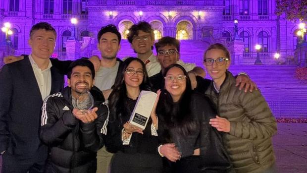 group of smiling students in front of purple lit building holding awards