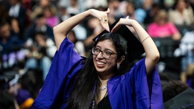 a graduate in a purple cap and gown making a heart gesture with her arms