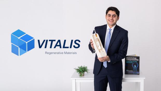 Student in suit holding product, posed next to company's logo featuring the words "Vitalis, Regenerative Materials"