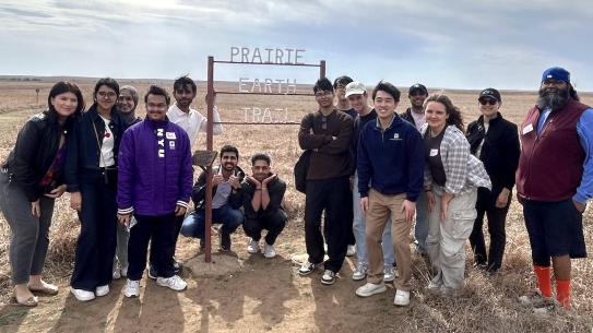 a group of students outside in an open prarie posing in front of a sign reading "Prairie Earth Trail"