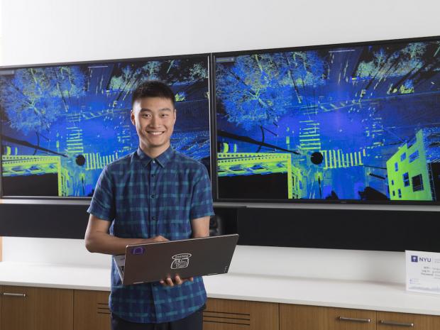 - smiling asian male student holding laptop in front of large digital screen