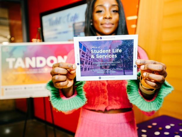 A student in colorful outfit holding a "student Life & Services" postcard