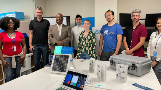 Eight people gathered around a square prototype, about the size of a large textbook.