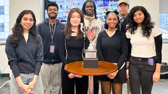 Seven students standing around a table with a trophy placed in the center. 